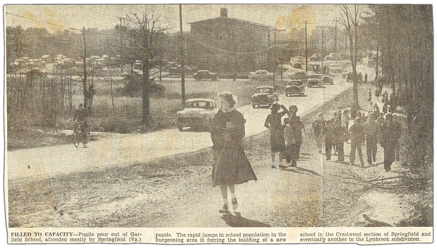 Black and white photograph from a newspaper clipping, published in The Evening Star on April 24, 1955. The caption reads: Filled to capacity. Pupils pour out of Garfield School, attended mostly by Springfield, Virginia pupils. The rapid jumps in school population in the burgeoning area is forcing the building of a new school in the Crestwood section of Springfield and eventually another in the Lynbrook subdivision. The picture was taken east of the school on Old Keene Mill Road. The road is barely wide enough to fit two cars. It was taken before all the shopping centers in this area were built and much of the surrounding area is still forest. Groups of children walk along a dirt path toward Backlick Road. One student is riding his bicycle up the street, followed by a long line of cars pulling out of the school’s driveway. 