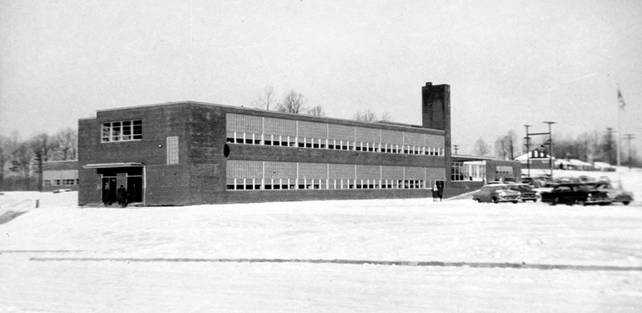 Black and white photograph of Crestwood Elementary School taken in 1958. There is snow on the ground. Cars are parked in the driveway in front of the school and four people are visible outside the building. The building is designed in the egg-crate style, common to schools of the 1950s, with rectangular classroom wings with rows of classrooms separated by a central hallway. The front classroom wing, office, and cafeteria are visible.  