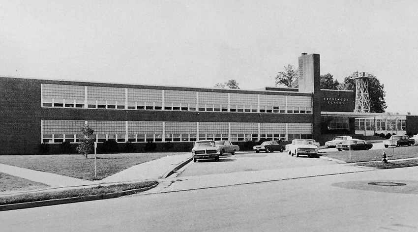 Black and white photograph of Crestwood Elementary School taken in 1965. The front of the building is pictured from Hanover Avenue. Cars are parked in what is now the bus loop in front of the building. A tall metal tower is visible behind the school.  